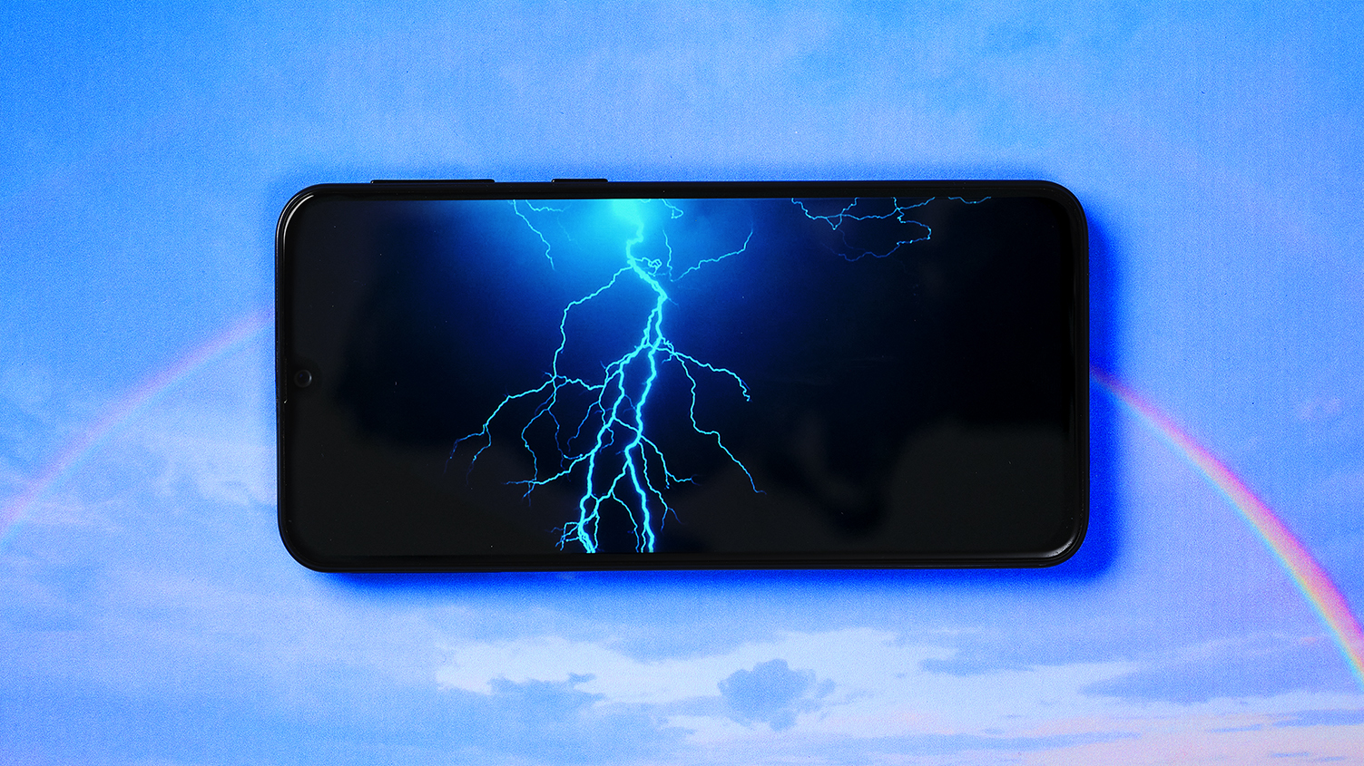A lightning storm on a mobile phone screen over the image of a rainbow in a blue sky.